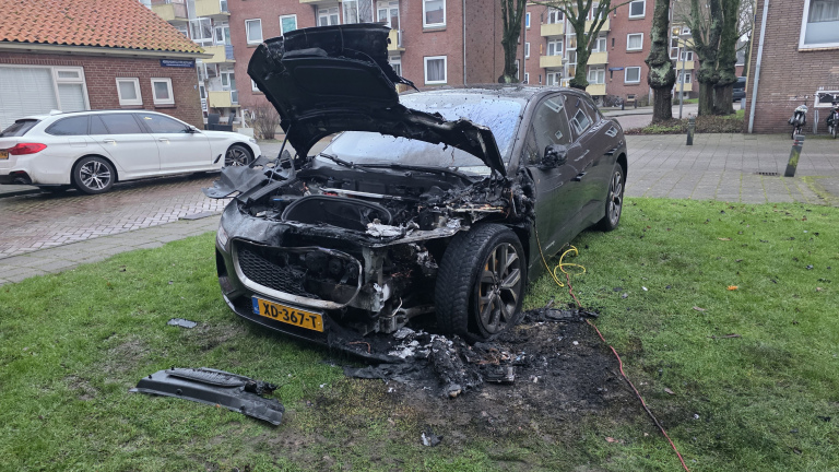 Auto met uitgebrand motorcompartiment en open motorkap op een grasveld, omringd door gebouwen en een geparkeerde witte auto.