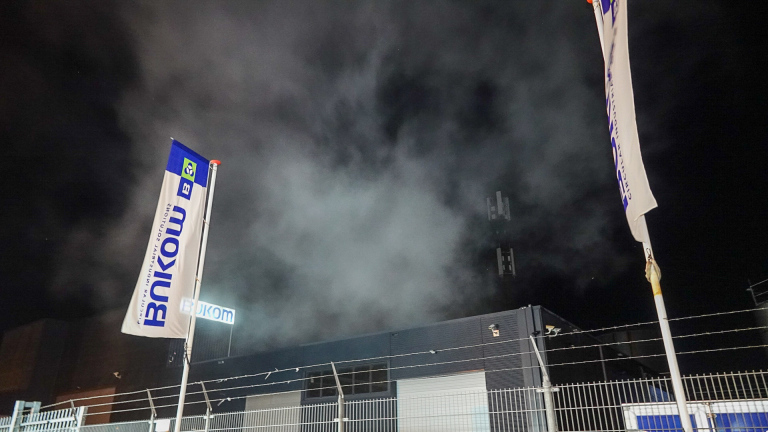 Industrieel gebouw bij nacht, met rookwolken en een zendmast zichtbaar tegen een donkere hemel.