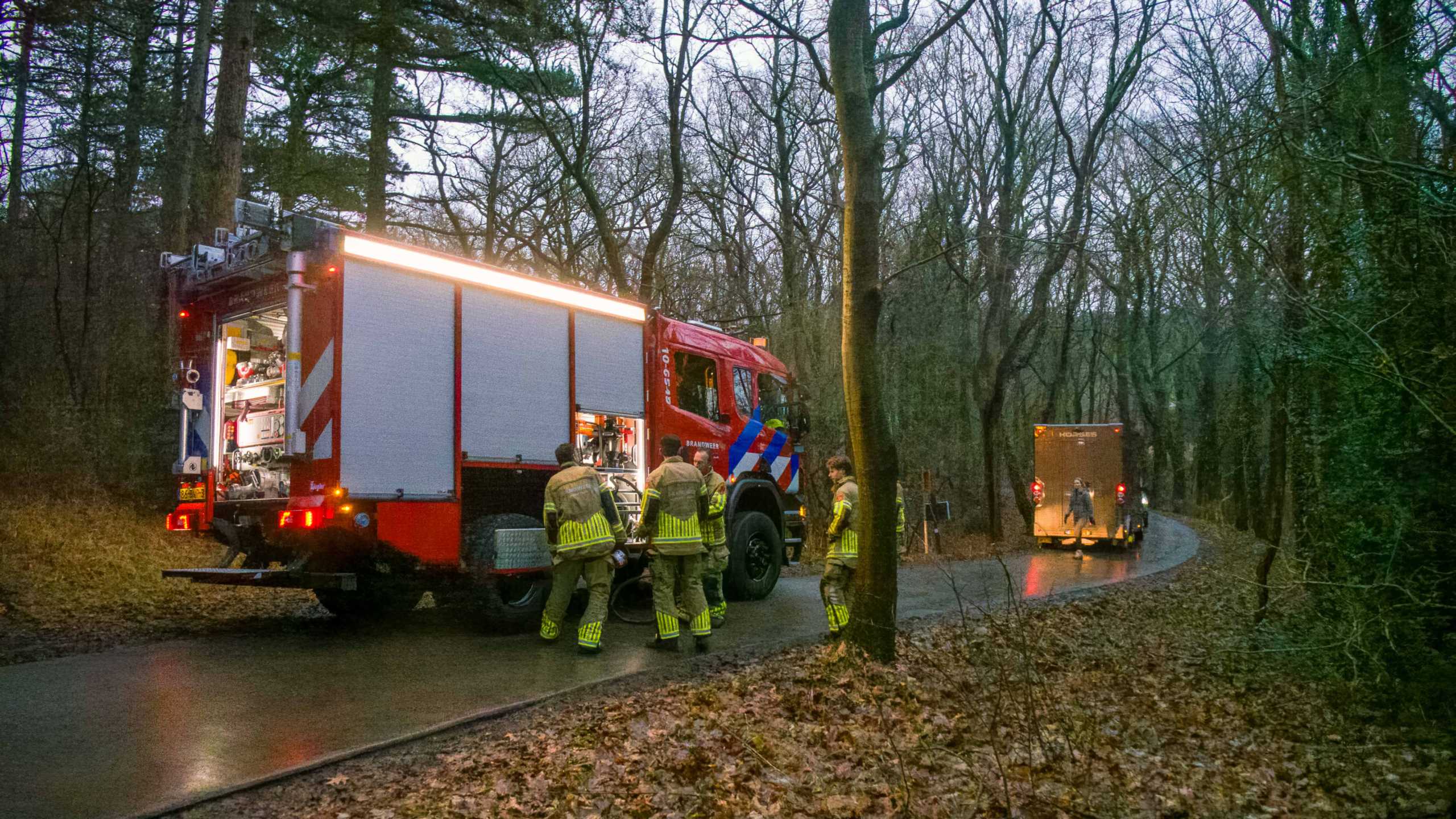 Brandweerlieden en mensen lopen op een modderig bospad omringd door dichte bomen.