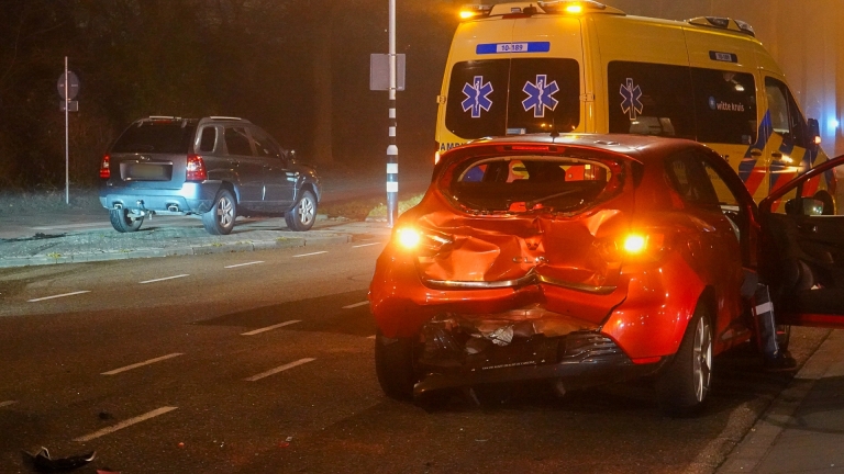 Twee ambulances en hulpverleners staan bij een rode auto met schade na een ongeval op een kruispunt 's nachts.
