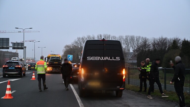 Gele onderhoudsvoertuigen met waarschuwingsborden op een drukke snelweg; een bushalte is zichtbaar aan de rechterkant.