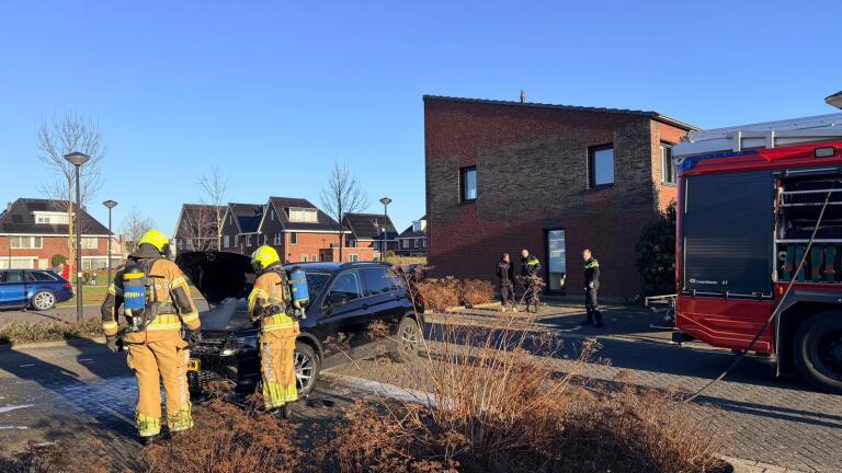 Brandweerlieden blussen een brand in de motorkap van een zwarte auto in een woonwijk.