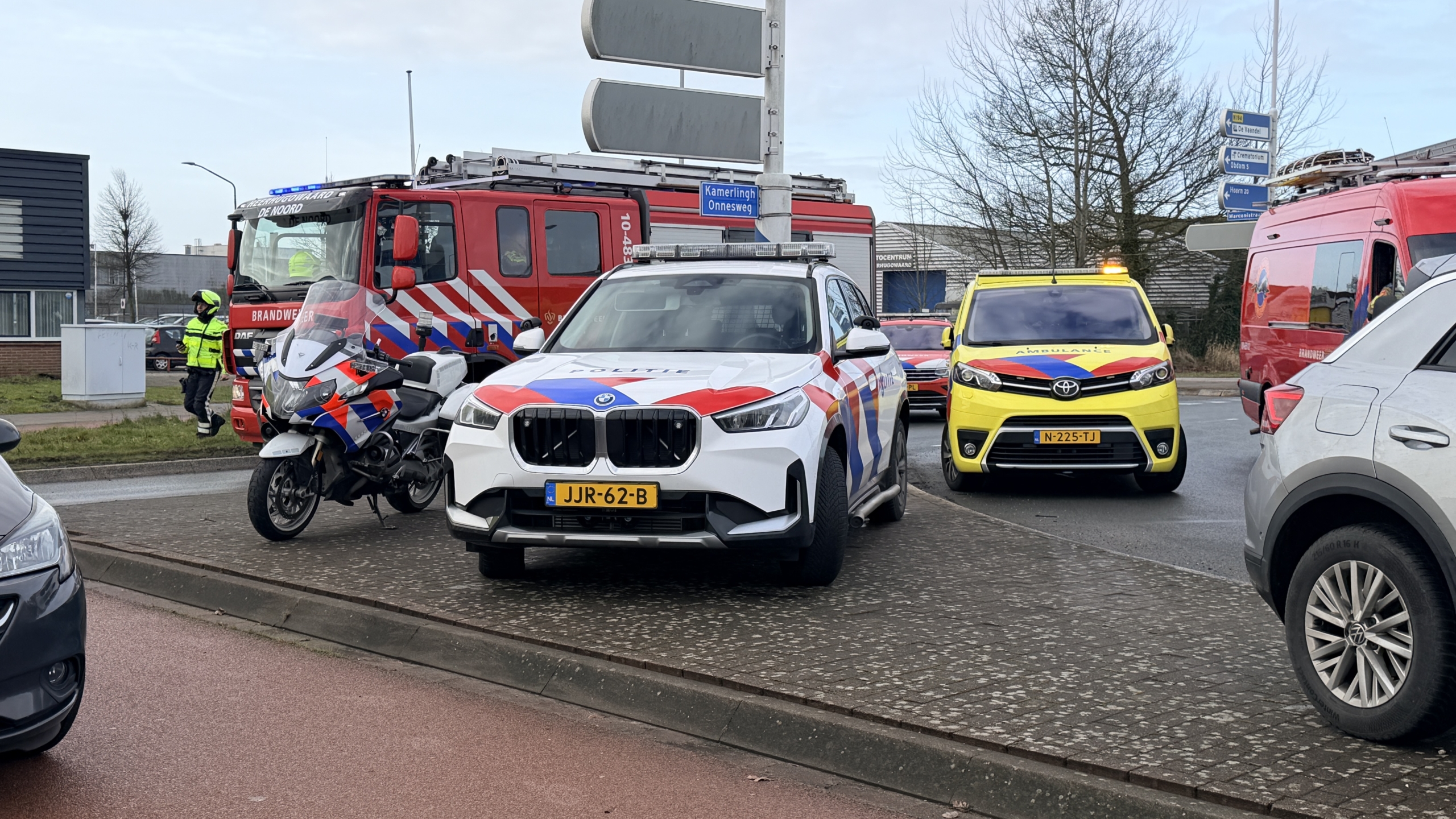 Auto half ondergedompeld in een sloot, met brandweer, ambulances en mensen op de achtergrond.