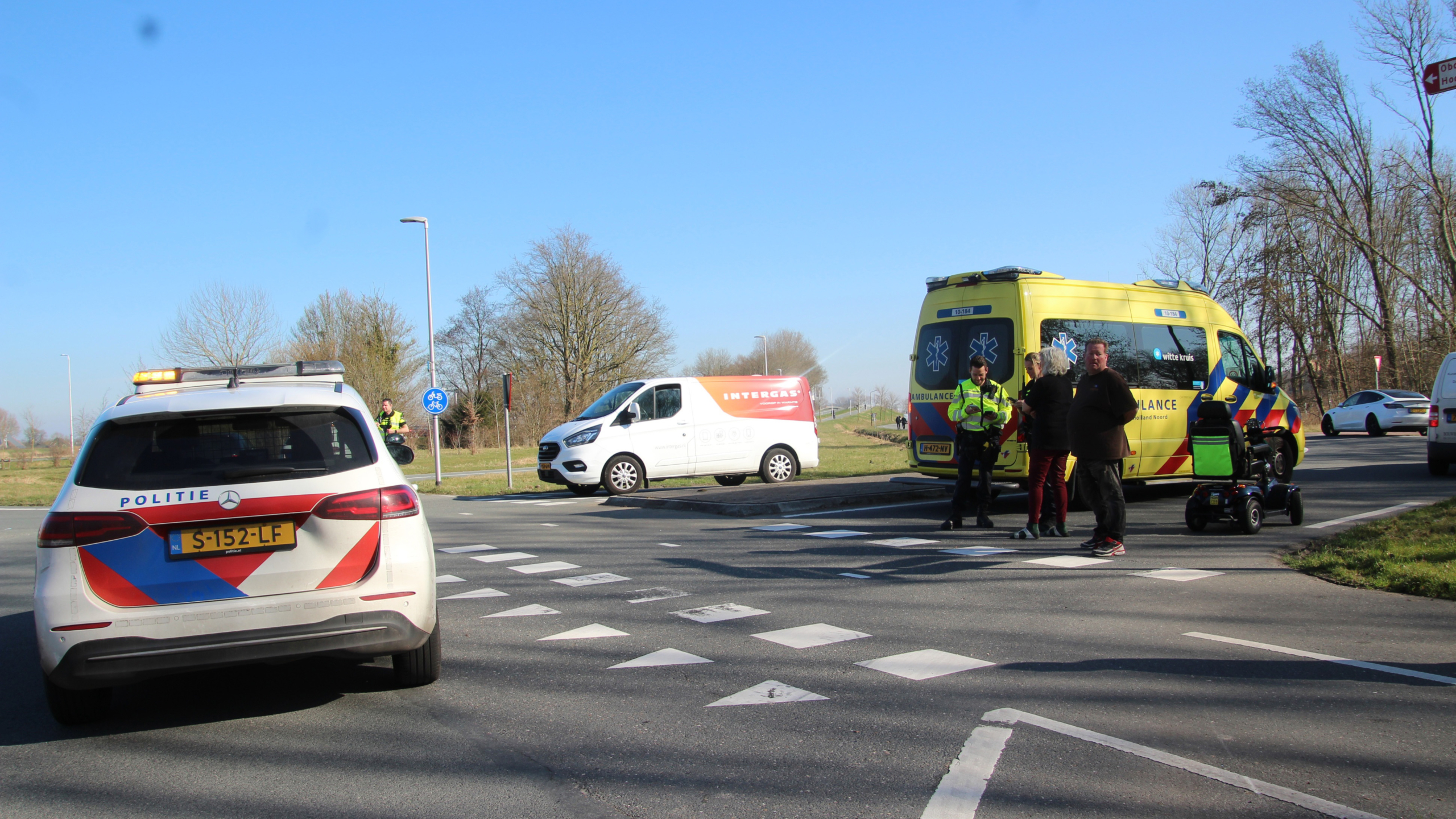 Scootmobiel op een kruispunt met politieagenten en een politiewagen op de achtergrond.