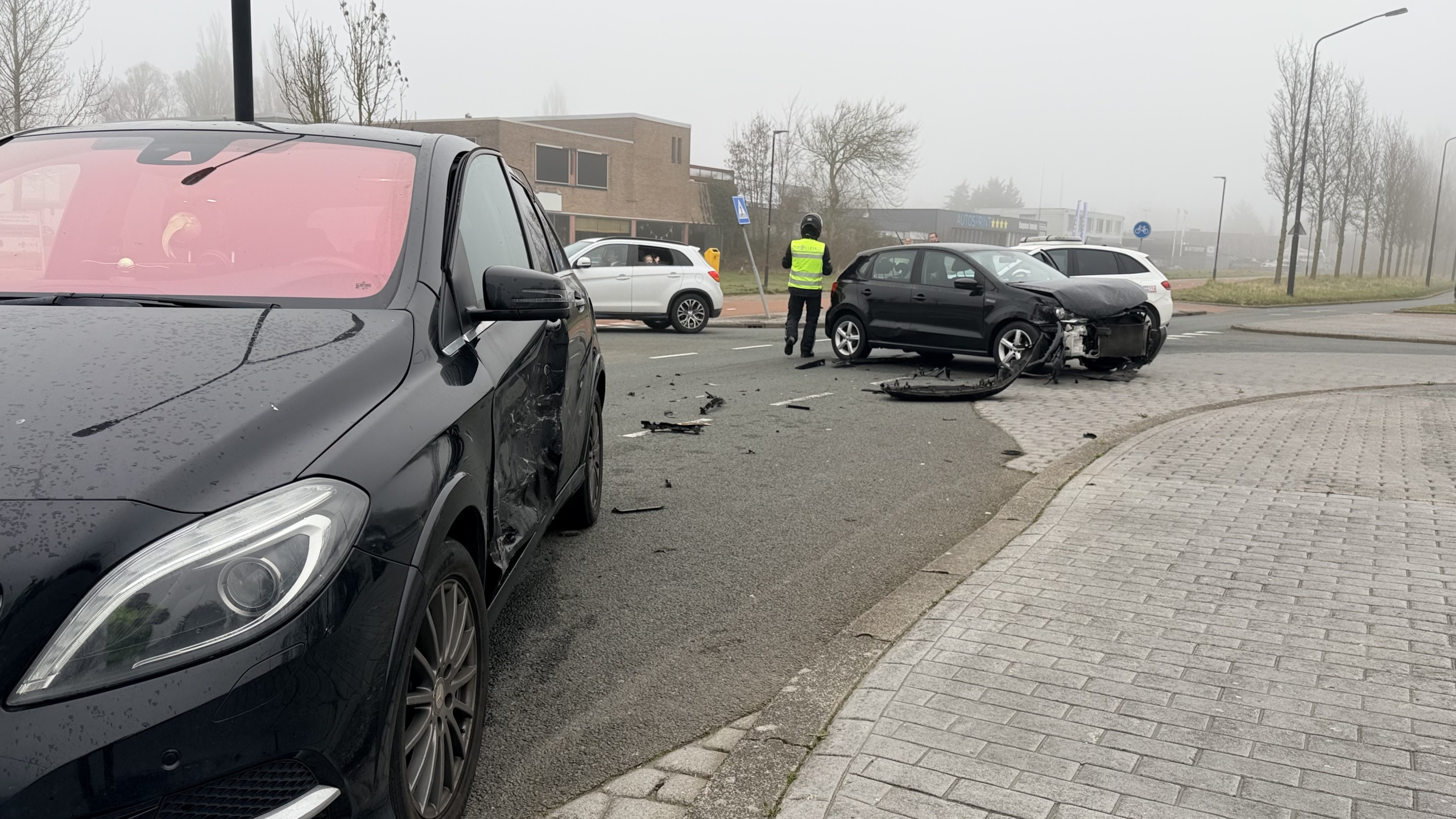 Auto met zware schade aan de voorzijde op een kruispunt, met politie en passerende voertuigen in de achtergrond.