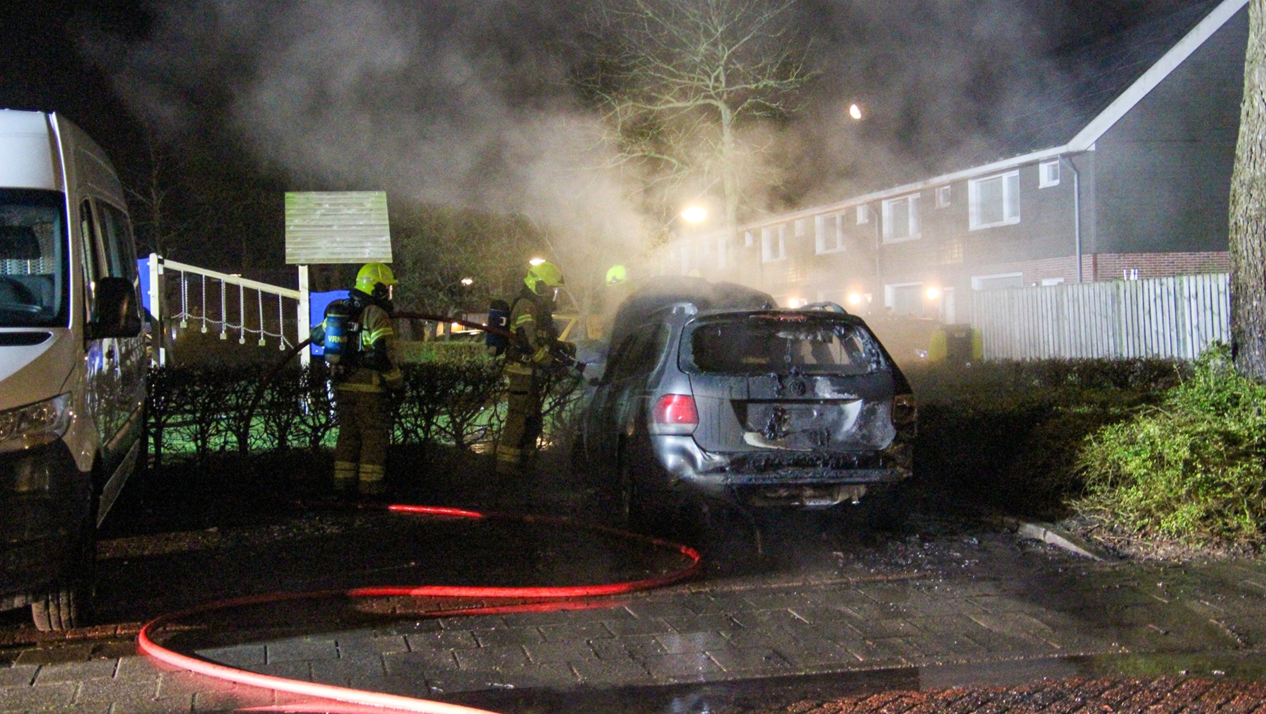 Auto in brand met grote vlammen, naast een boom en een andere geparkeerde auto in een nachtelijke omgeving.
