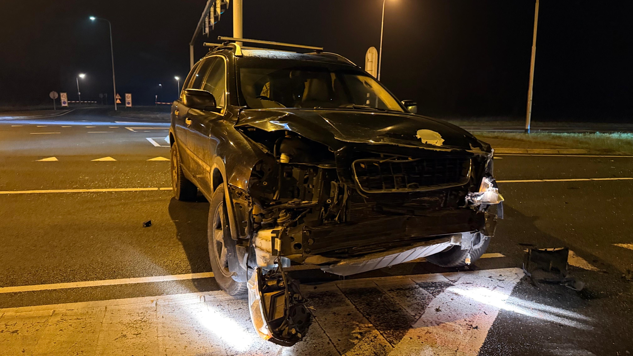 Auto met deukschade en geopende airbags bij een rotonde 's nachts, met verzamelde mensen en kijkers op de achtergrond.