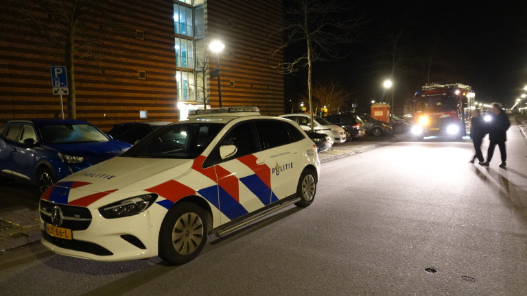 Brandweertruck en brandweerlieden bij een gebouw in de nacht, met geparkeerde auto’s op de voorgrond.