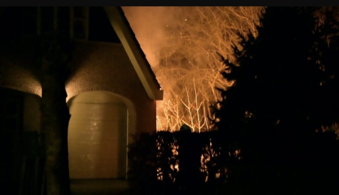 Een nachtelijk tafereel van een huis met rook die uit het dak komt, en een paar mensen die buiten staan.