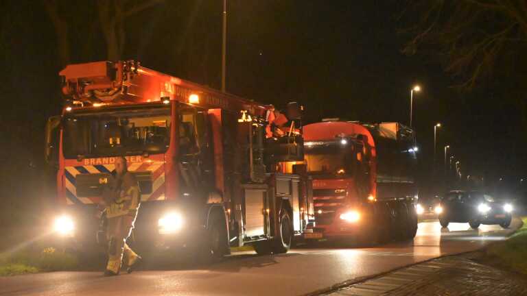 Een nachtelijk tafereel van een huis met rook die uit het dak komt, en een paar mensen die buiten staan.