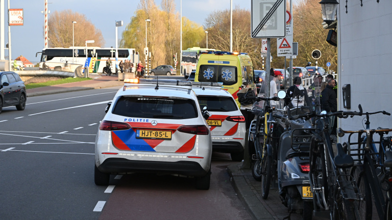 Politieagenten voeren een gesprek op een trottoir naast een geparkeerde politiewagen, omringd door voorbijgangers en fietser.