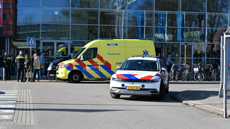 Een politieauto en een ambulance staan geparkeerd voor een gebouw met glazen gevel, omringd door enkele mensen en fietsen.