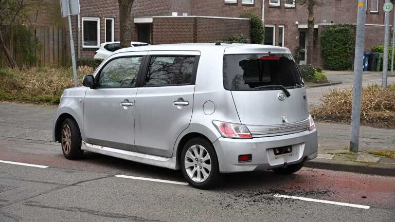 Zilverkleurige auto met een gebroken achterruit geparkeerd langs een rustige straat tegenover een huis.