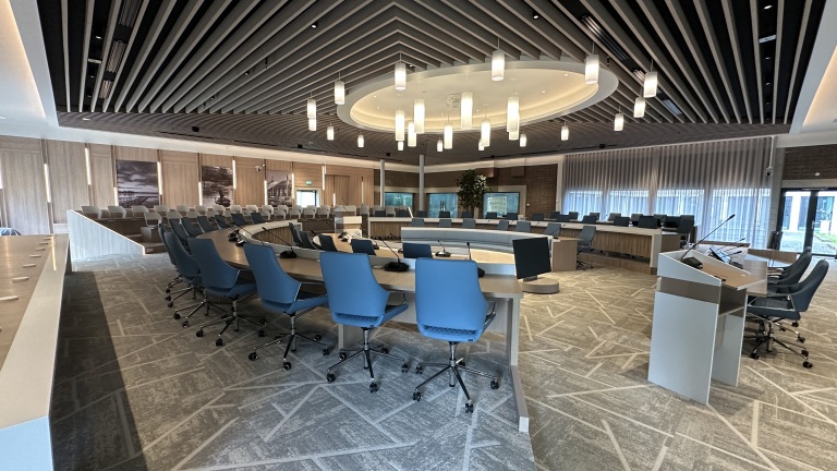 Vergaderzaal met rondom een grote ovale tafel, blauwe stoelen en moderne hanglampen aan het plafond.