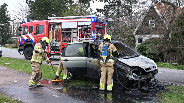Brandweerlieden blussen een uitgebrande auto langs de weg met een brandweerwagen op de achtergrond.