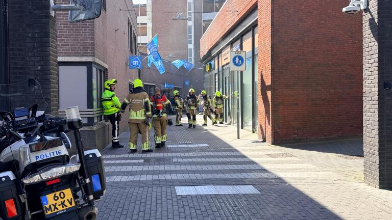 Brandweerlieden en politie bij een winkelstraat, met een politieagent op een motor en Albert Heijn vlaggen zichtbaar.