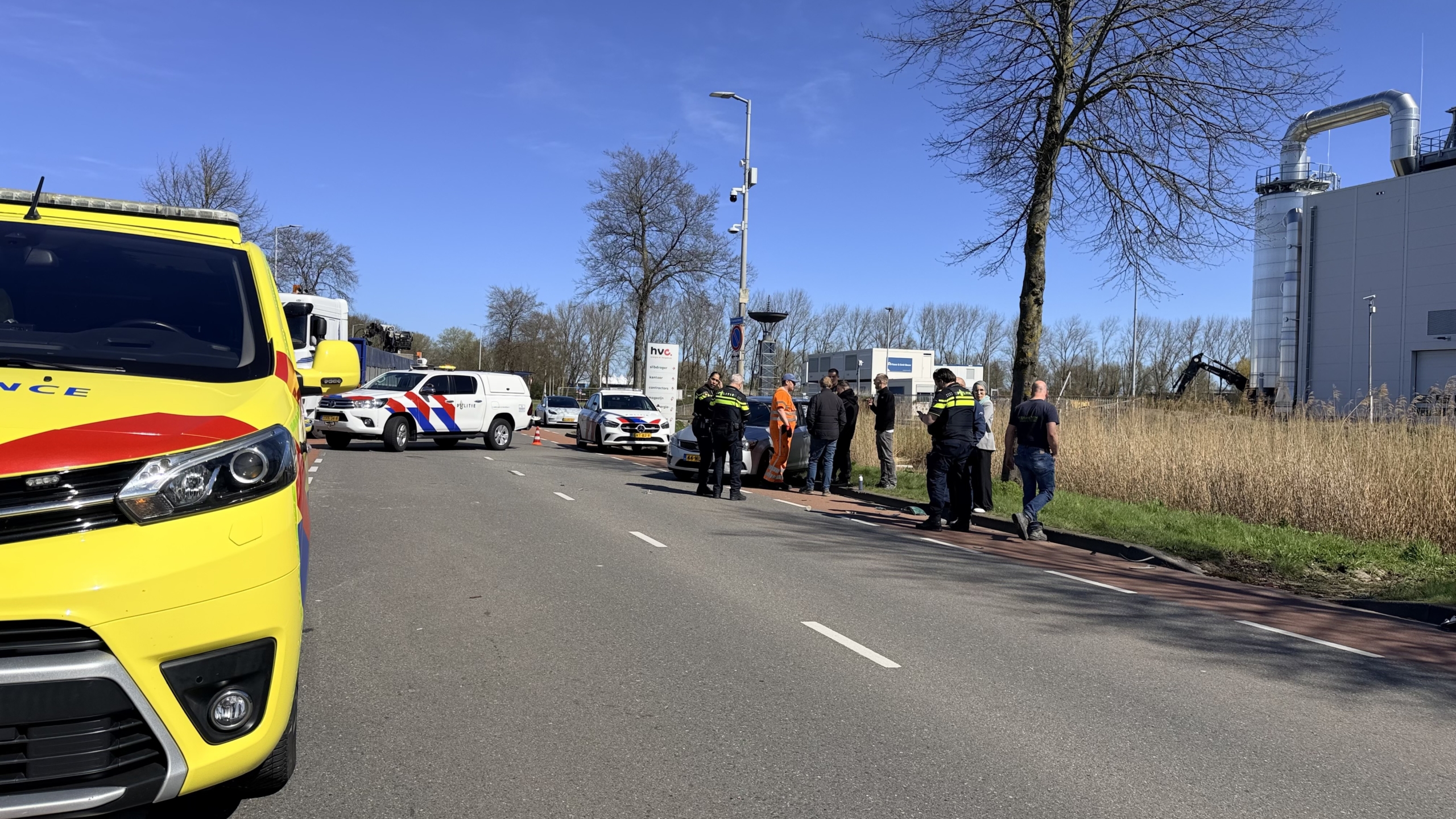 Een zilveren auto met zware zijkantschade staat naast de weg; een politieagent en een persoon in oranje veiligheidskleding zijn erbij aanwezig.