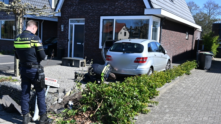 Een zilverkleurige auto is tegen een bakstenen muur en een tuinhek gebotst, met schade aan de bumper.
