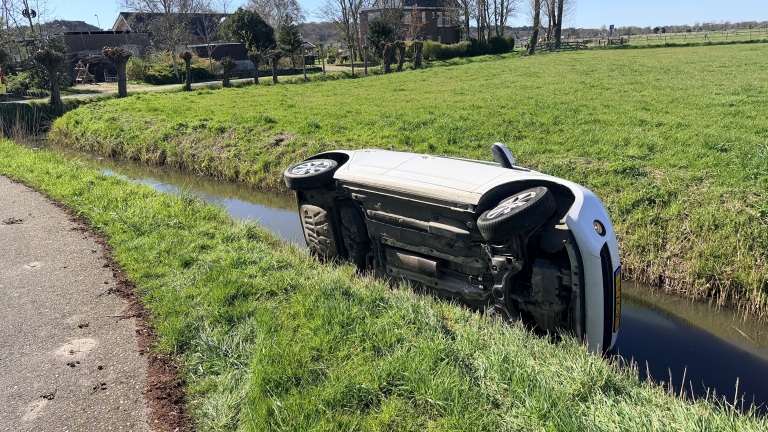 Auto op zijn kant in een sloot naast een grasveld en een weg in een landelijke omgeving.