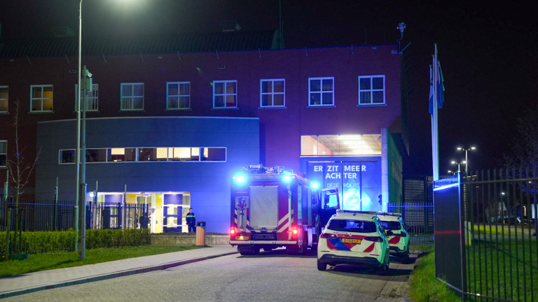 Brandweer- en politievoertuigen met zwaailichten voor een gebouw 's nachts, met zichtbaar personeel bij de ingang.