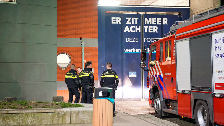 Brandweer- en politievoertuigen met zwaailichten voor een gebouw 's nachts, met zichtbaar personeel bij de ingang.