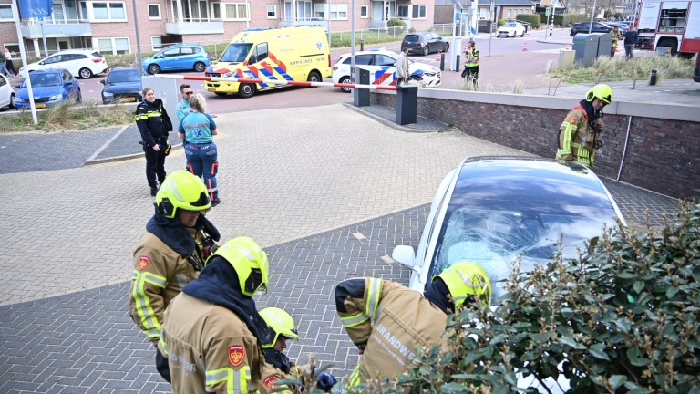 Brandweerlieden en politie bij een auto-ongeluk met een beschadigd voertuig; ambulance en politieagenten op de achtergrond.