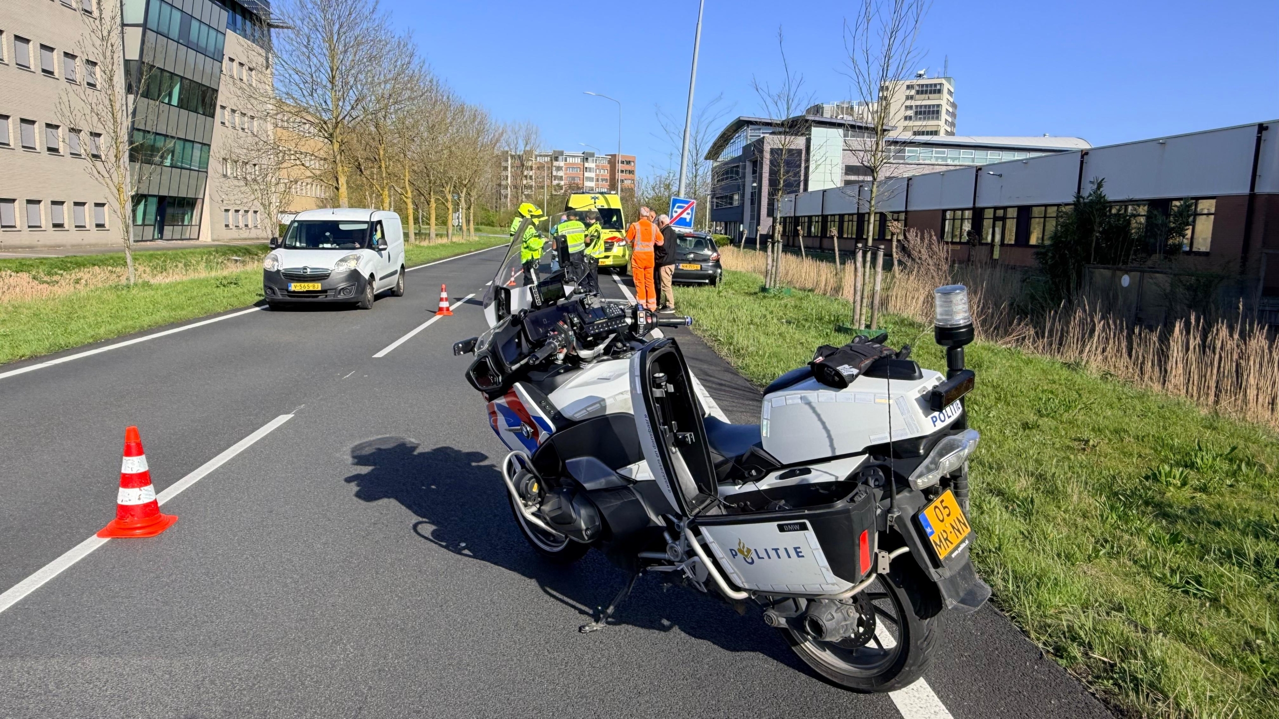 Auto met schade na botsing tegen een paal langs een weg, met verkeerskegel en mensen op de achtergrond.