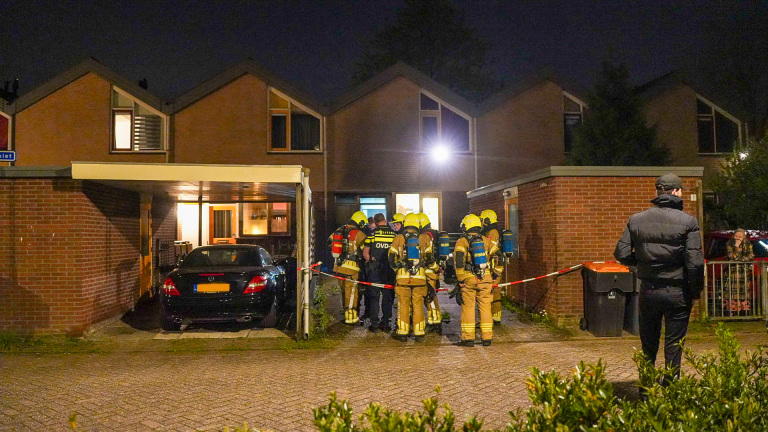 Politieagenten praten buiten een huis terwijl brandweerlieden naar binnen gaan, met een zwarte auto op de oprit.