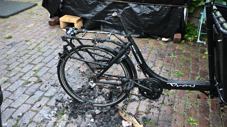Verbrande fiets op een natte bakstenen straat, met zichtbare schade aan het frame en de wielen.