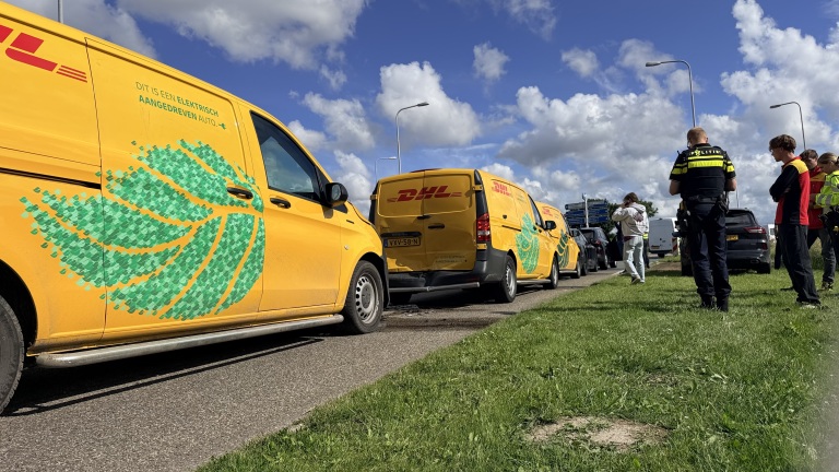 Gele DHL-bestelwagens met groene patronen staan langs de weg; politie en mensen kijken toe.