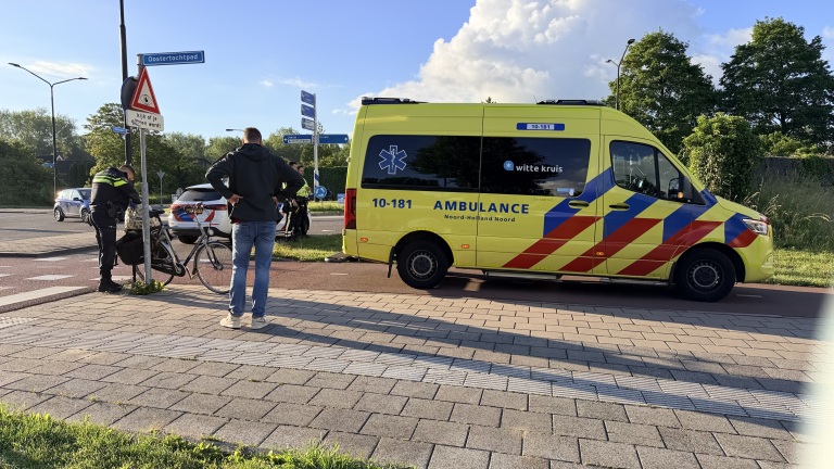 Een gele ambulance staat geparkeerd langs de weg bij een kruispunt, met politieagenten en een fiets in de buurt.