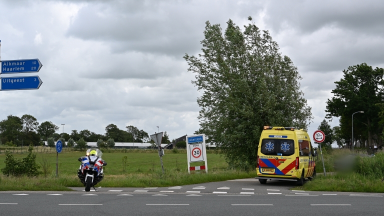 Een ambulance en een politieagent op een motor bij de ingang van Akersloot, omringd door groen landschap.
