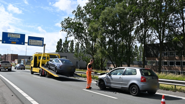 Auto-ongeluk op de snelweg met een beschadigde zilverkleurige auto en een gele bergingswagen die een zwarte auto vervoert.