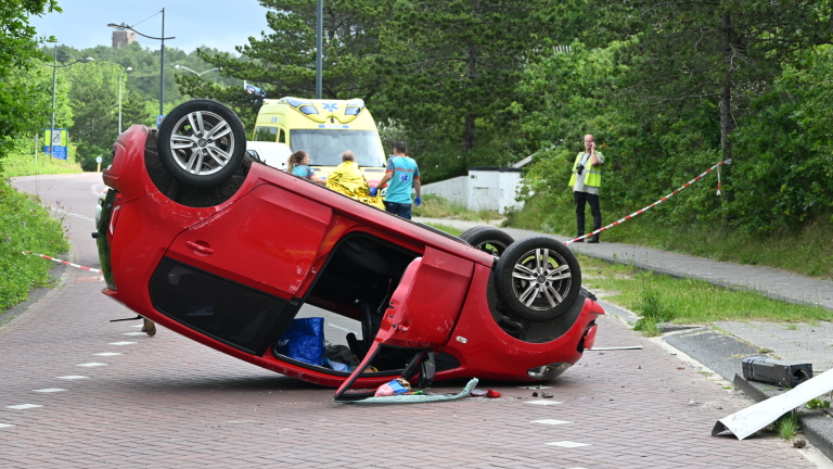 Een rode auto ligt omgekeerd op de weg, met een ambulance en hulpverleners op de achtergrond.