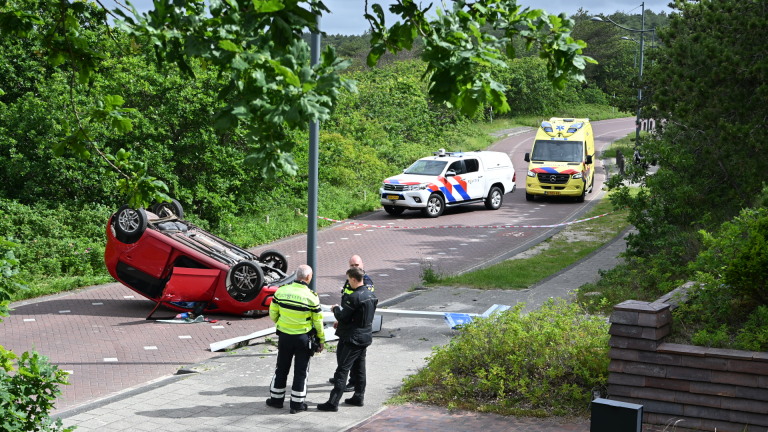 Een rode auto ligt omgekeerd op de weg, met een ambulance en hulpverleners op de achtergrond.
