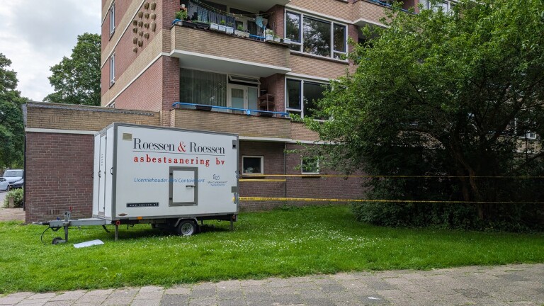 Wit bedrijfstrailer van Roessen & Roessen asbestsanering geparkeerd bij een appartementsgebouw, omgeven door waarschuwingslint.