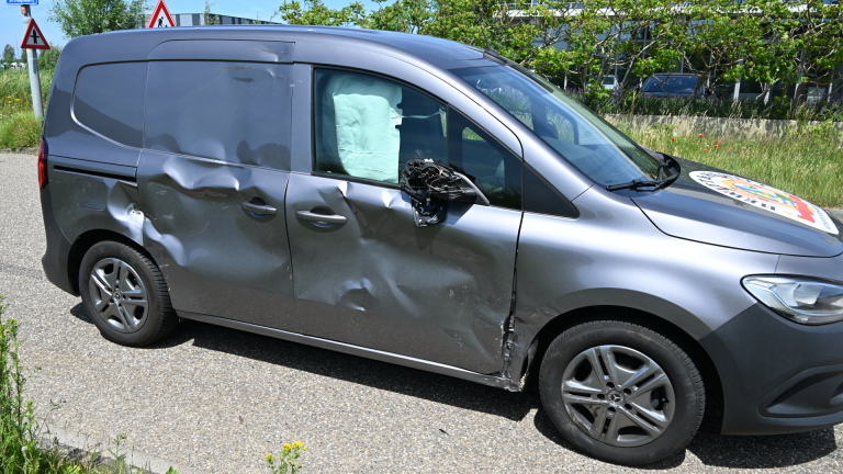 Grijze bestelwagen met aanzienlijke deuken aan de zijkant en een kapotte zijspiegel, geparkeerd op een weg.