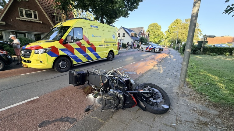 Omgevallen motorfiets op de stoep naast een ambulance en politieauto, met omstanders aan de kant van de weg.
