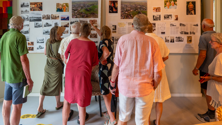 Een groep oudere mensen bekijkt fotocollages op een tentoonstellingswand in een galerij.