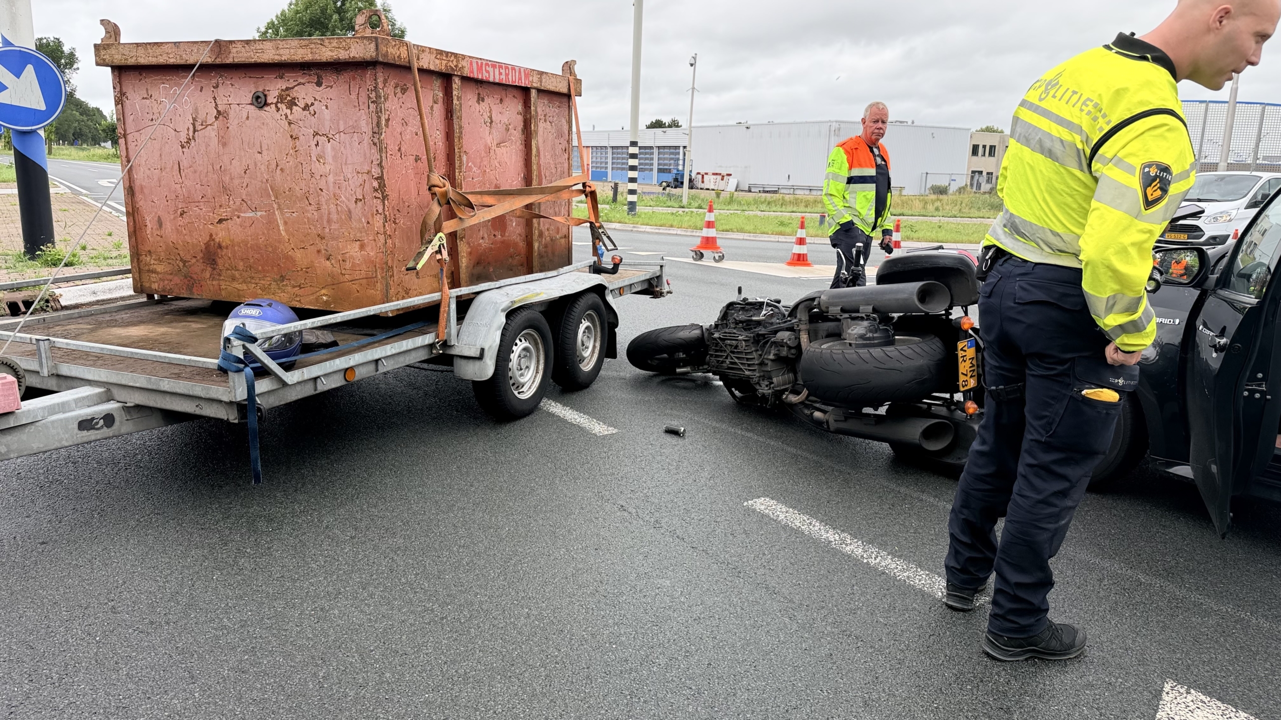 Omgevallen motorfiets op de grond naast een auto en aanhanger op een kruispunt, met verkeer en verkeerskegels op de achtergrond.