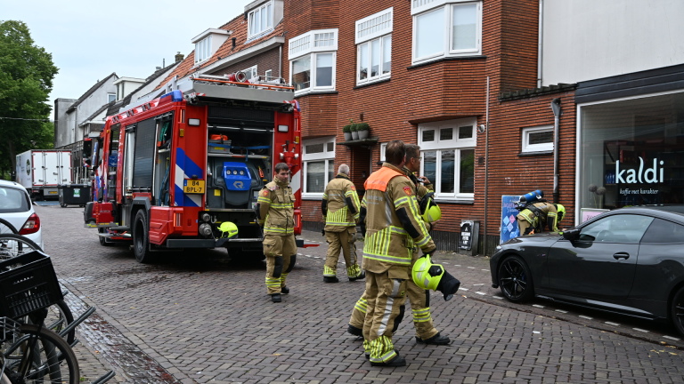 Brandweerauto met open achterklep en brandweerlieden op een straat bij bakstenen panden en winkel Kaldi