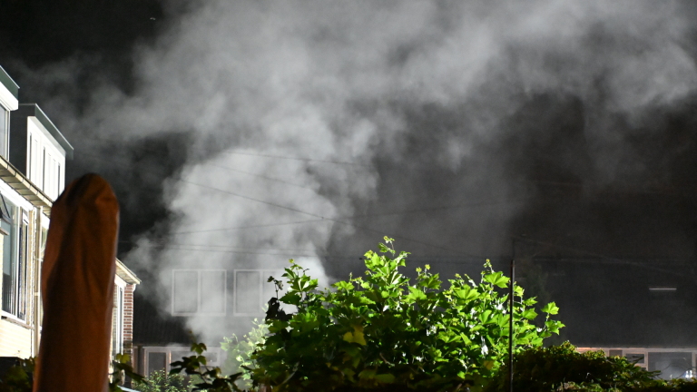 Dikke rookwolken stijgen ’s nachts op achter huizen, met fel verlichte groene struiken op de voorgrond