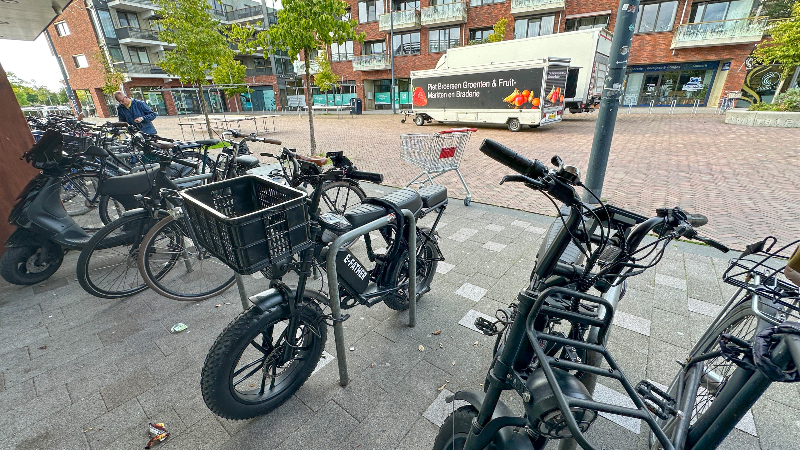 Geparkeerde fatbike met grote zwarte krat voorop, naast andere fietsen op een plein bij appartementen en winkelwagen