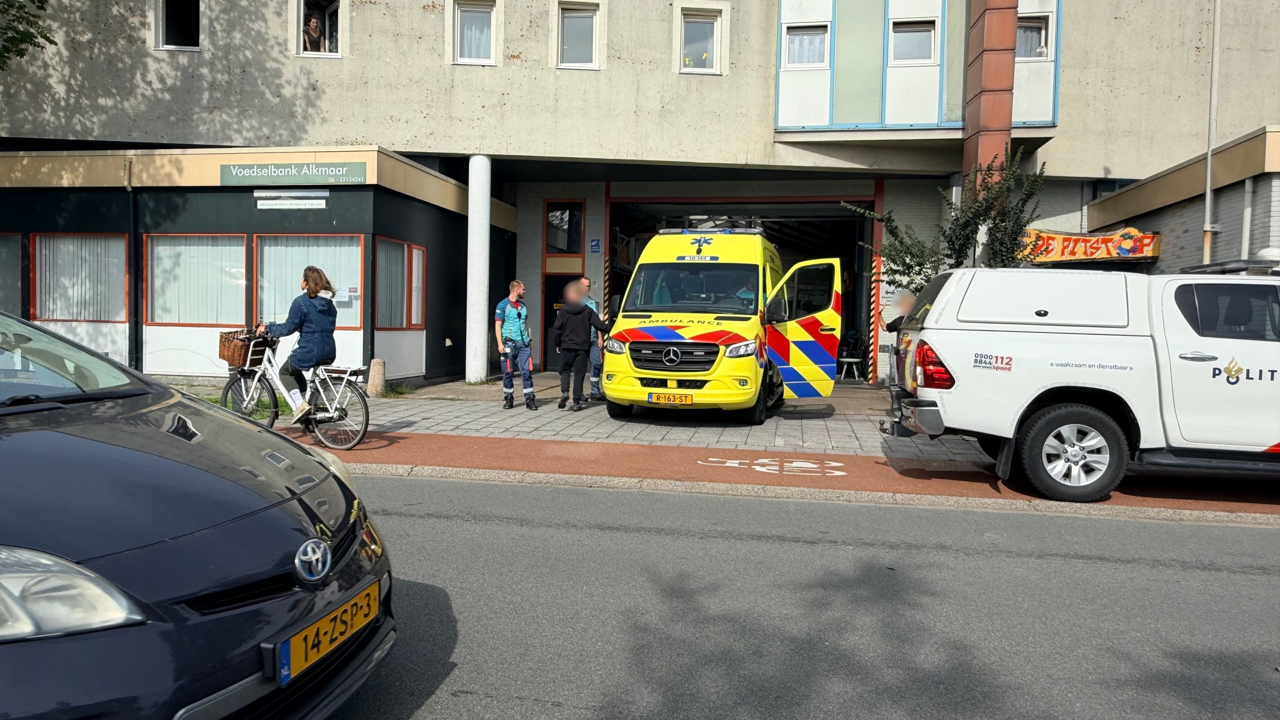 Ambulance en politieauto’s staan langs de weg bij een gebouw, met hulpverlener en omstanders op de stoep