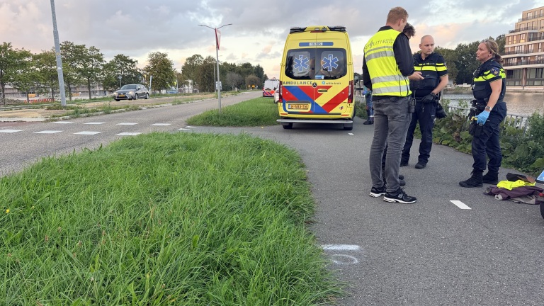 Politieagenten praten naast een geparkeerde ambulance op een fietspad langs het water, met kleding op de grond