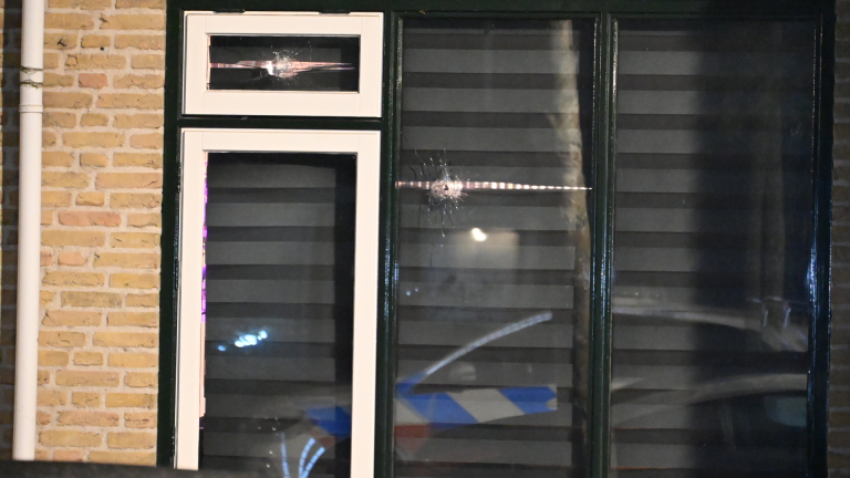 Ruit van woning met jaloezieën en twee kogelgaten in het glas, naast bakstenen muur en raamkozijnen