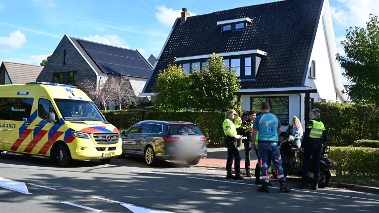 Ambulance en politie bij een stilstaande auto; hulpverleners praten met een vrouw naast een scooter in woonwijk